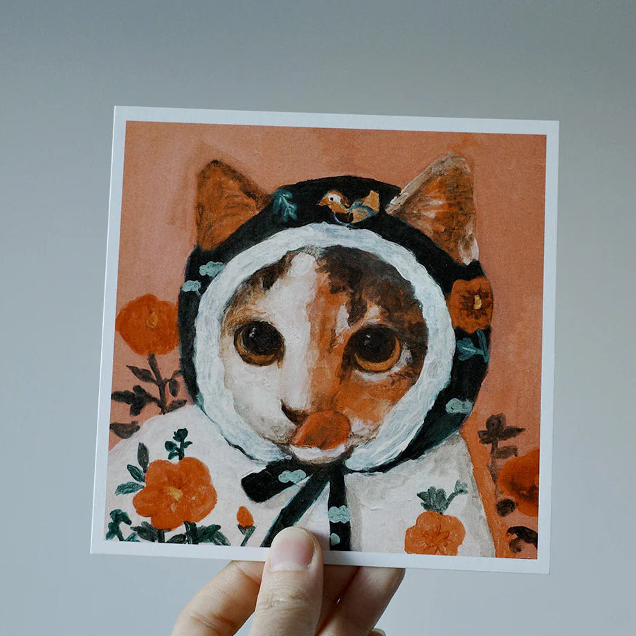 Illustration of a cat wearing a hood with floral patterns, held against a plain background.