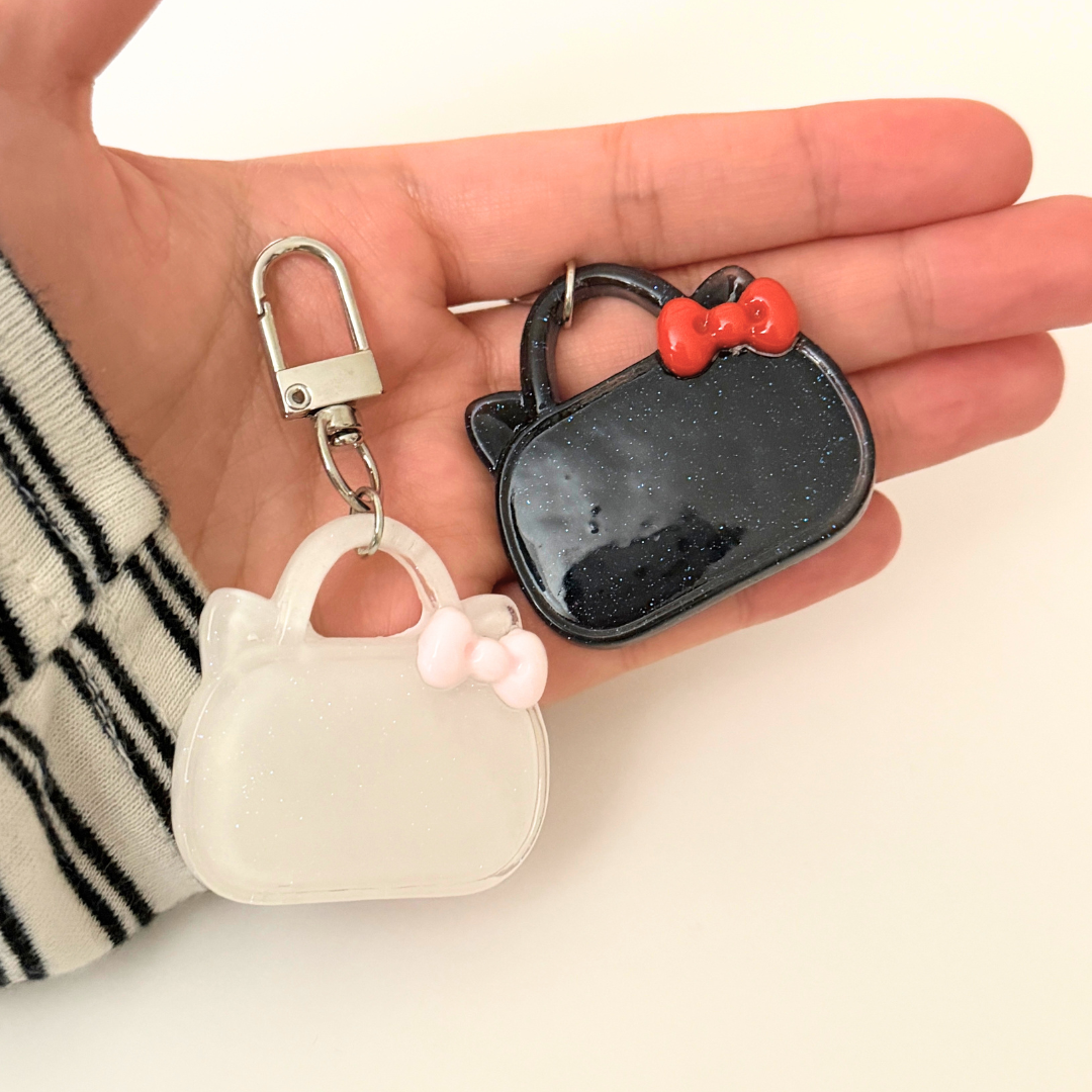 Two small handbag-shaped keychains, one black with a red bow and one white, held in a hand against a light background.