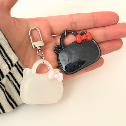 Two small handbag-shaped keychains, one black with a red bow and one white, held in a hand against a light background.