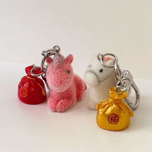 Set of keychains including a red bag, pink plush animal, and gold bag on a white background